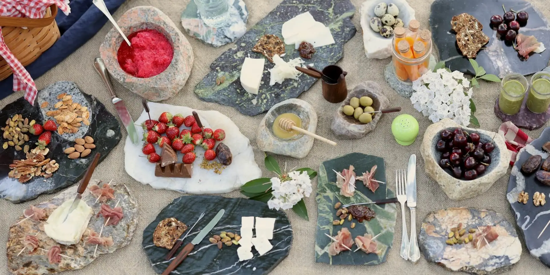 Picnic spread on natural stone serving boards and bowls, featuring cheese, cured meats, fruit, nuts, honey, and drinks.