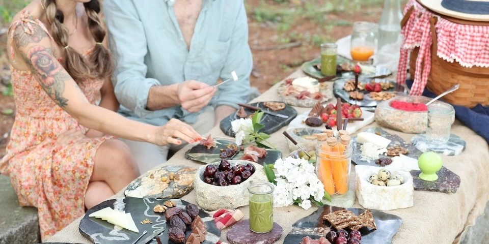 wo people enjoying an outdoor picnic with cheese, cured meats, fruit, and drinks served on natural stone boards and bowls.
